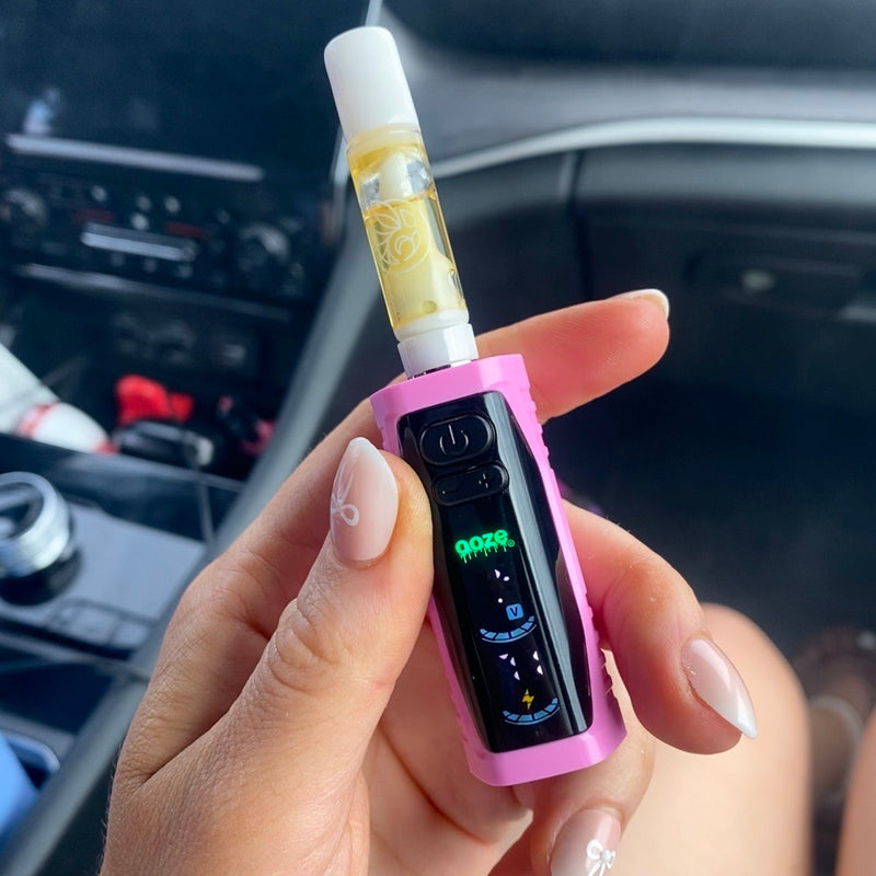 Close up of a hand holding a pink Ooze Runt Mini battery with a white ceramic cartridge connected and the screen turned on, while sitting in the passenger seat of a car