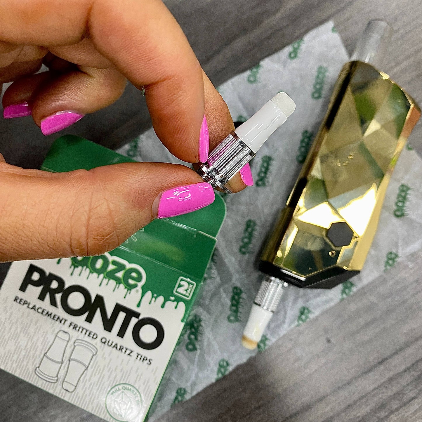 A girl with pink nails is holding a Pronto quartz replacement coil tip above a gold pronto and the coil box that are laying on a table