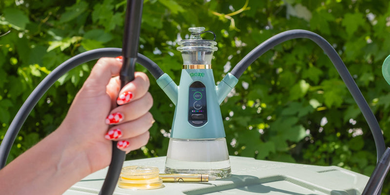 A mint Ooze Ripley dab rig hookah set on an outdoor table, with a hand holding the black hose in the foreground against a background of green foliage.