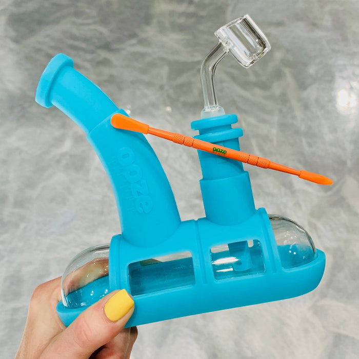 Holding up the aqua teal Ooze steamboat silicone bong with the quartz banger inserted to use as a dab rig. An orange metal dab tool is resting on the magnet on the side of the downstem