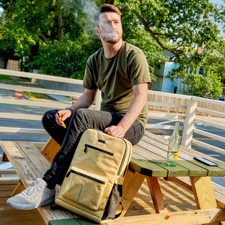 Man smoking outdoors with a backpack and bottle on a wooden table