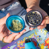 Opening a teal saturn grinder to show the ground bud in the chamber, held above a rolling tray