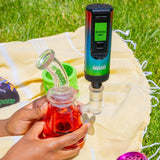 A rainbow Ooze Verge dry herb vape is turned on and inserted into a red Glyco freeze bong with the water pipe adapter. The photo is taken outside on a yellow blanket on the grass