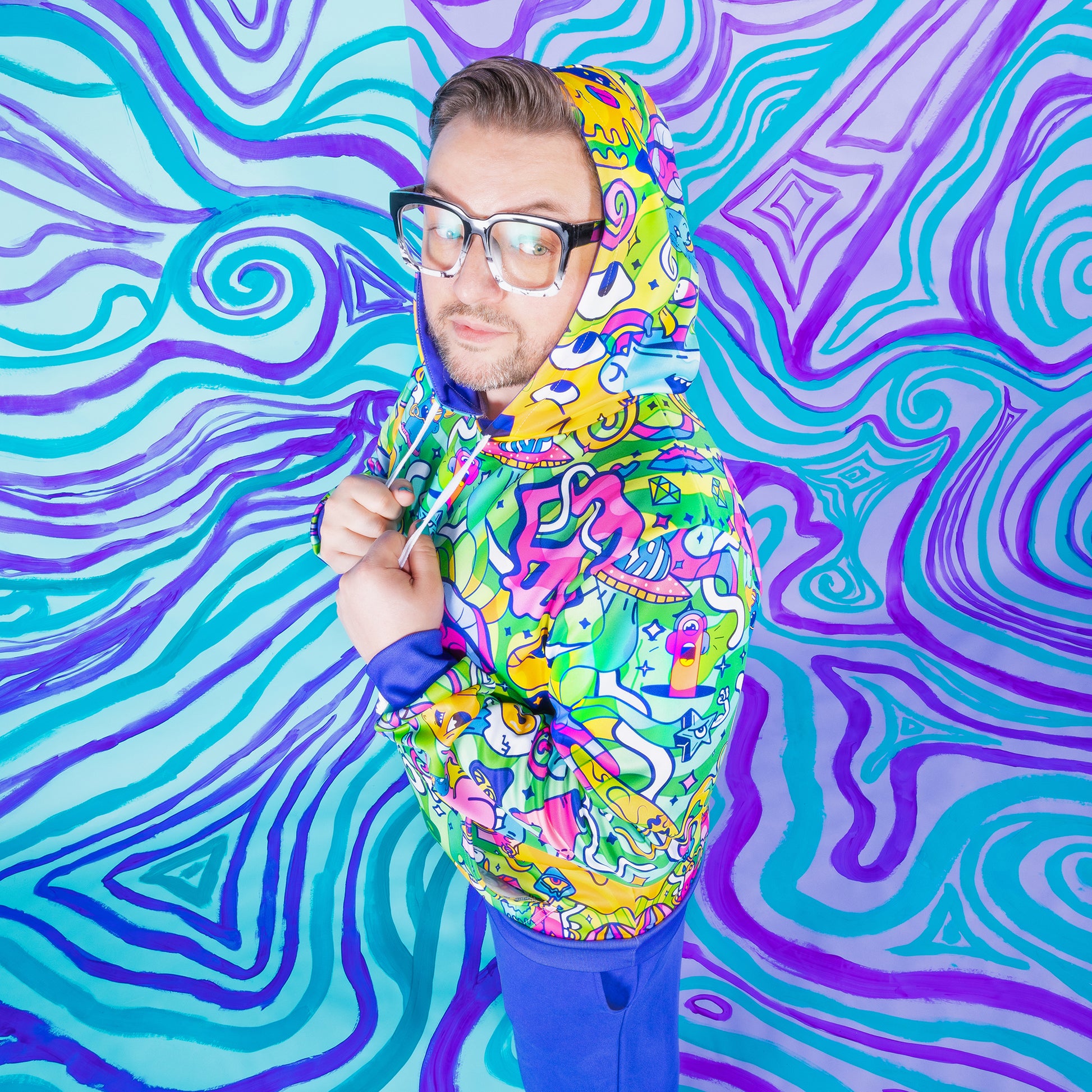 A white guy with square chunky glasses is wearing a Chroma hoodie with the hood on. He's standing on an angle so you can see more of the design, and tugging on both strings.