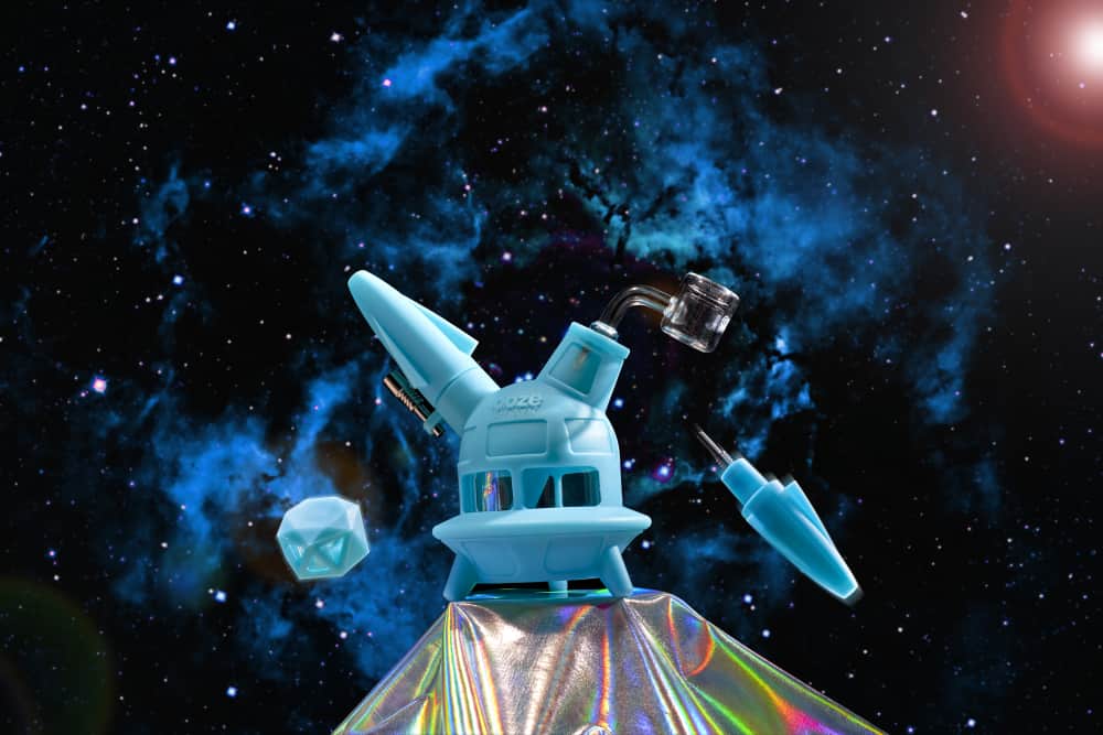 The aqua teal Ooze UFO is shown as a dab rig with the banger inserted. It has a galaxy background and the dab straw and stash jar and floating