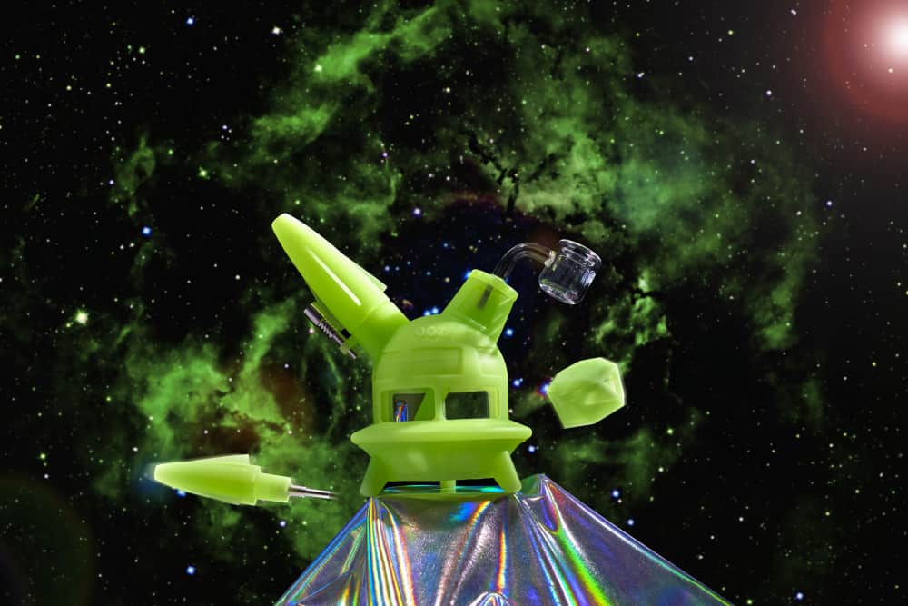 The green glow Ooze UFO is shown as a dab rig against a galaxy background with the dab straw and stash jar floating next to it